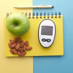 Flat lay of diabetes tools, green apple, almonds, and notebook for health planning.